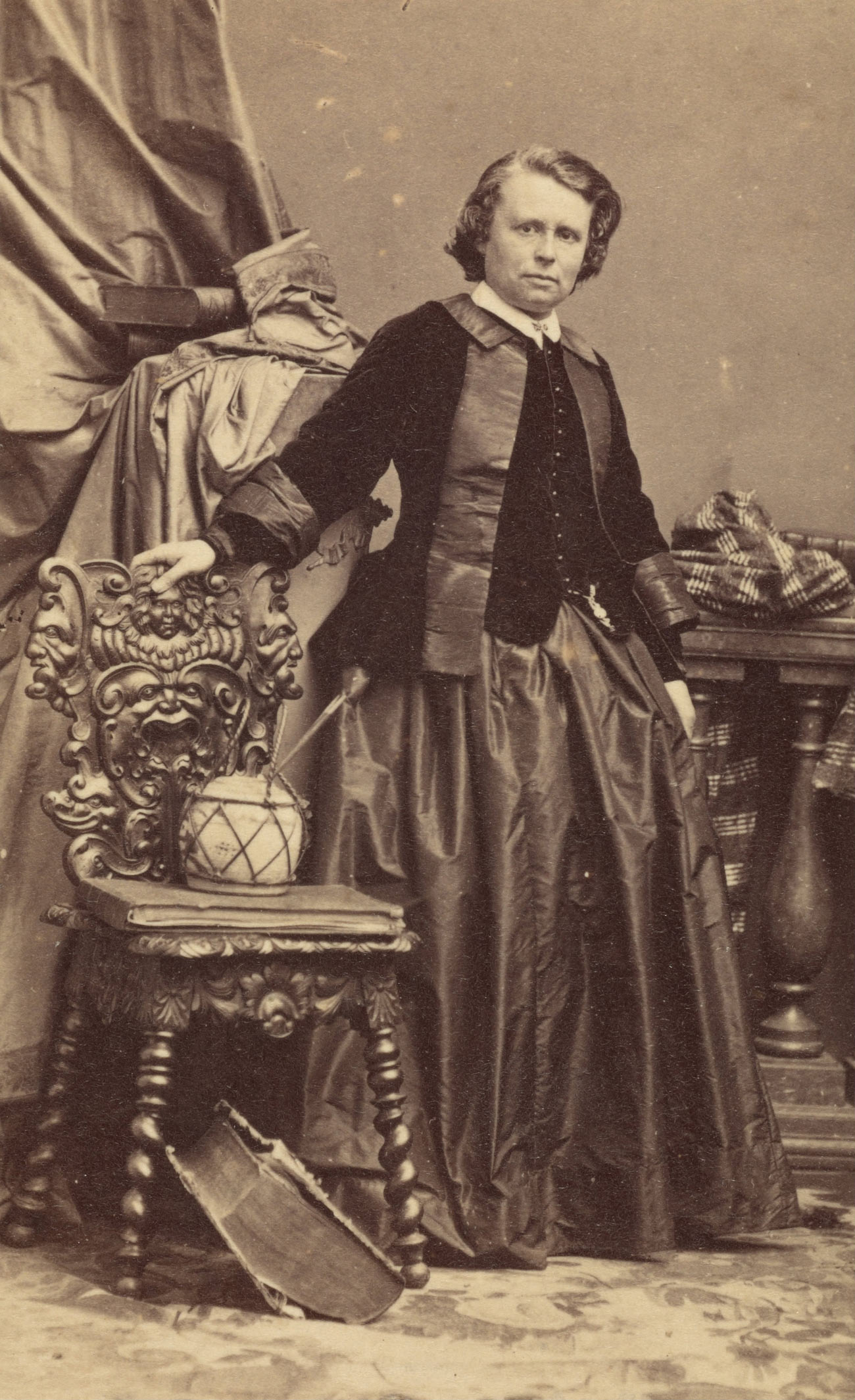 Rosa Bonheur wears a velvet jacket over a long skirt. She stands facing the camera with her right hand on the back of an elaborately carved chair.
