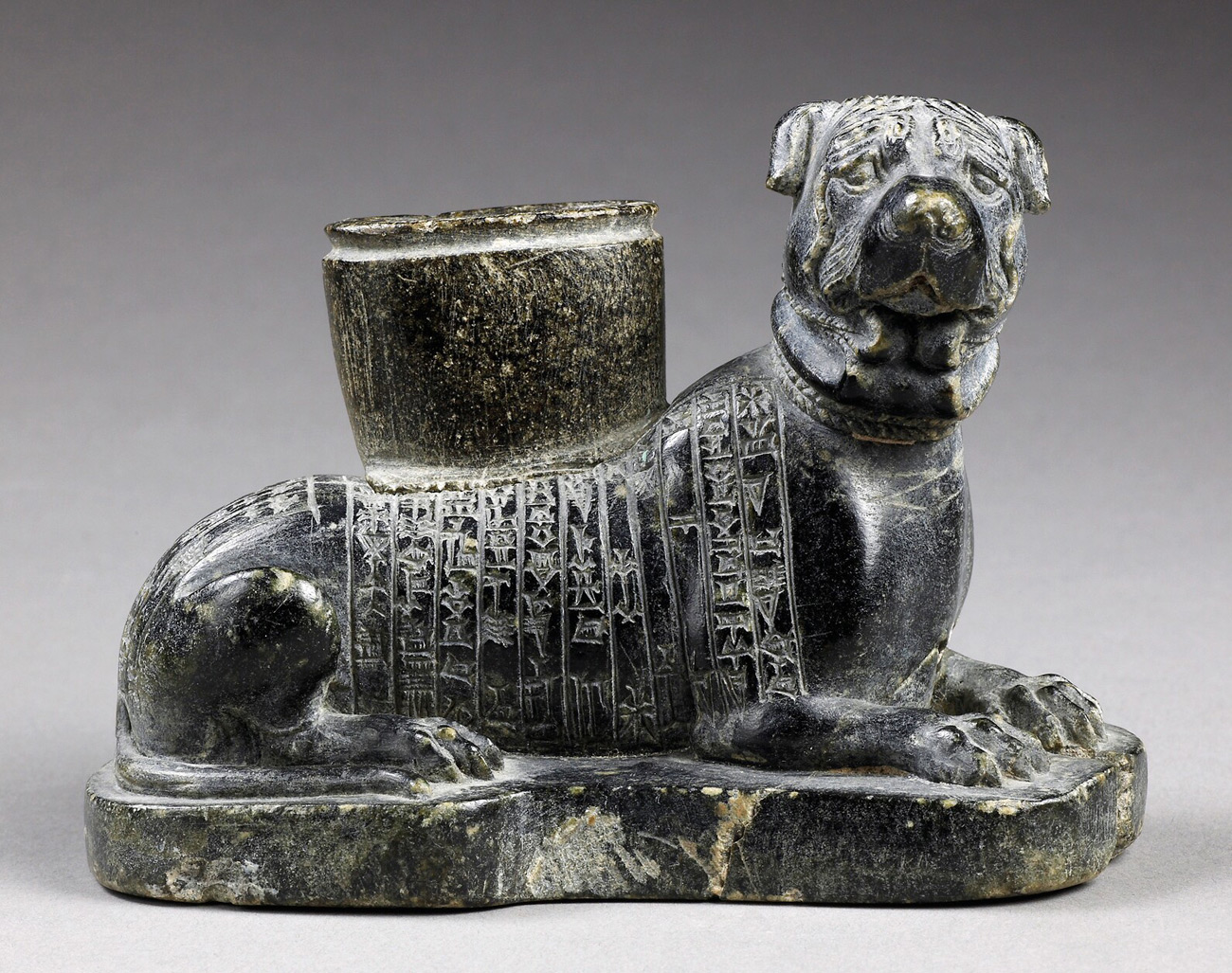 Dark-colored statue of a dog laying down with its head up, with symbols engraved in its side and a small round holder on its back