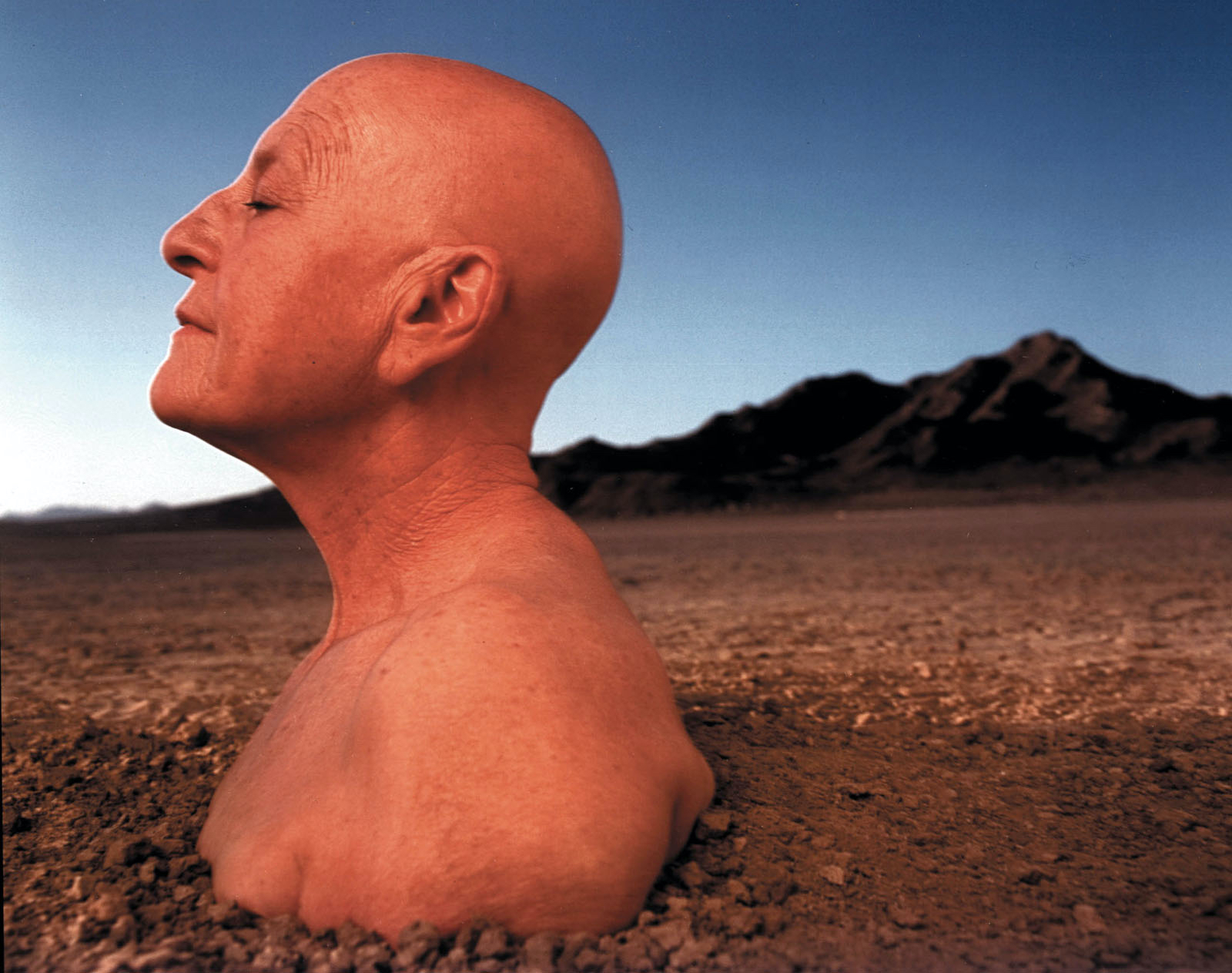 a giant bald head and torso rise from a red desert landscape