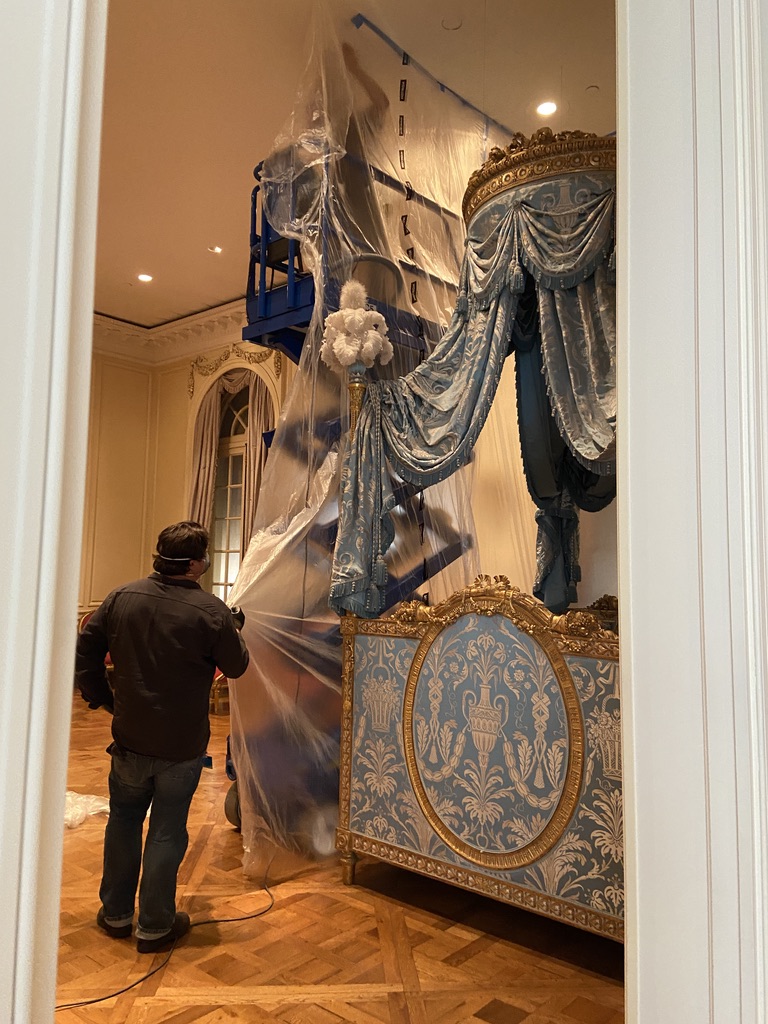 An ornate blue and gold canopy bed is wrapped in plastic while a staff man looks on