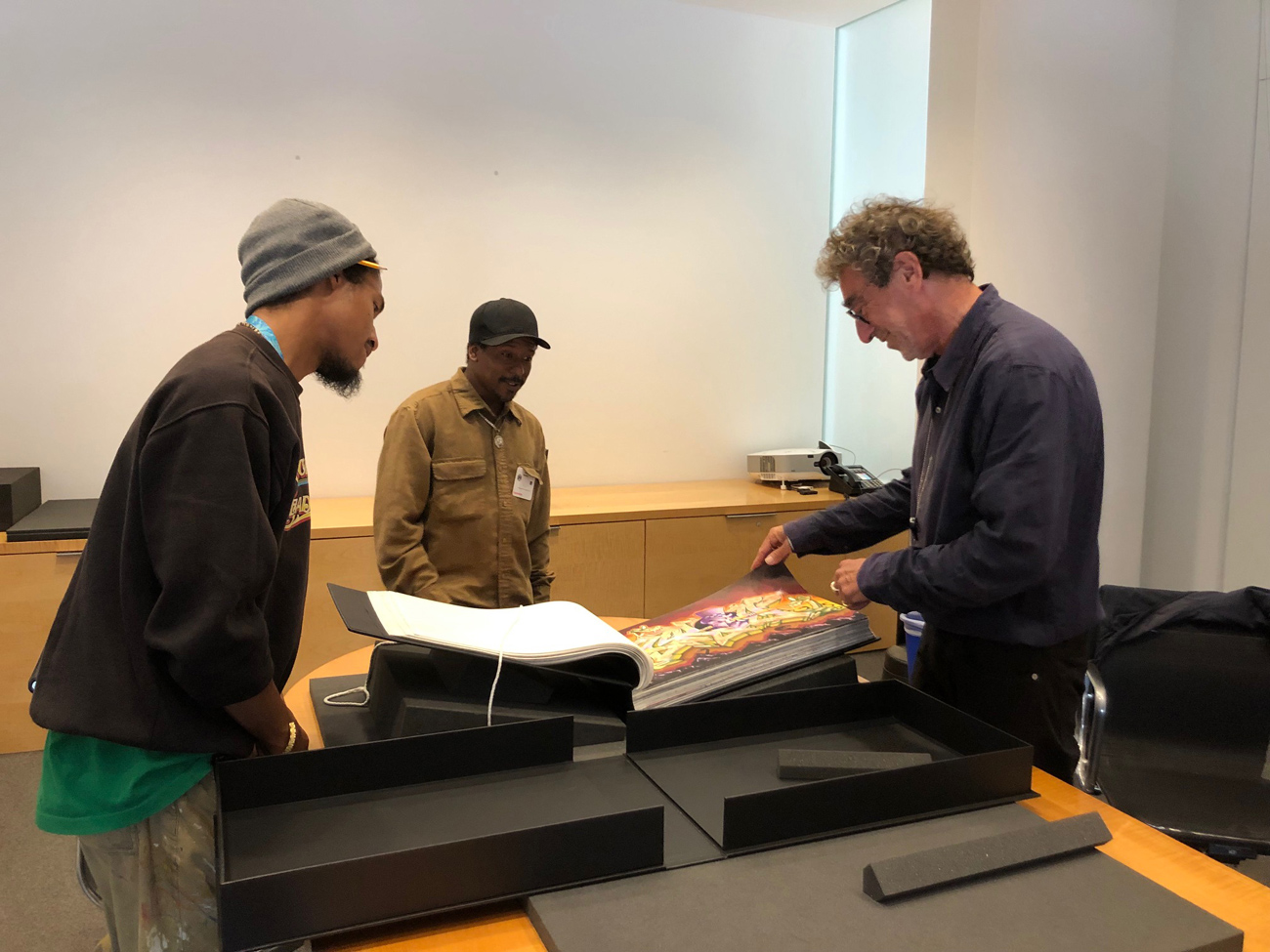 Three men stand in a conference room, looking at a large black book sitting on a pedestal on a long table.
