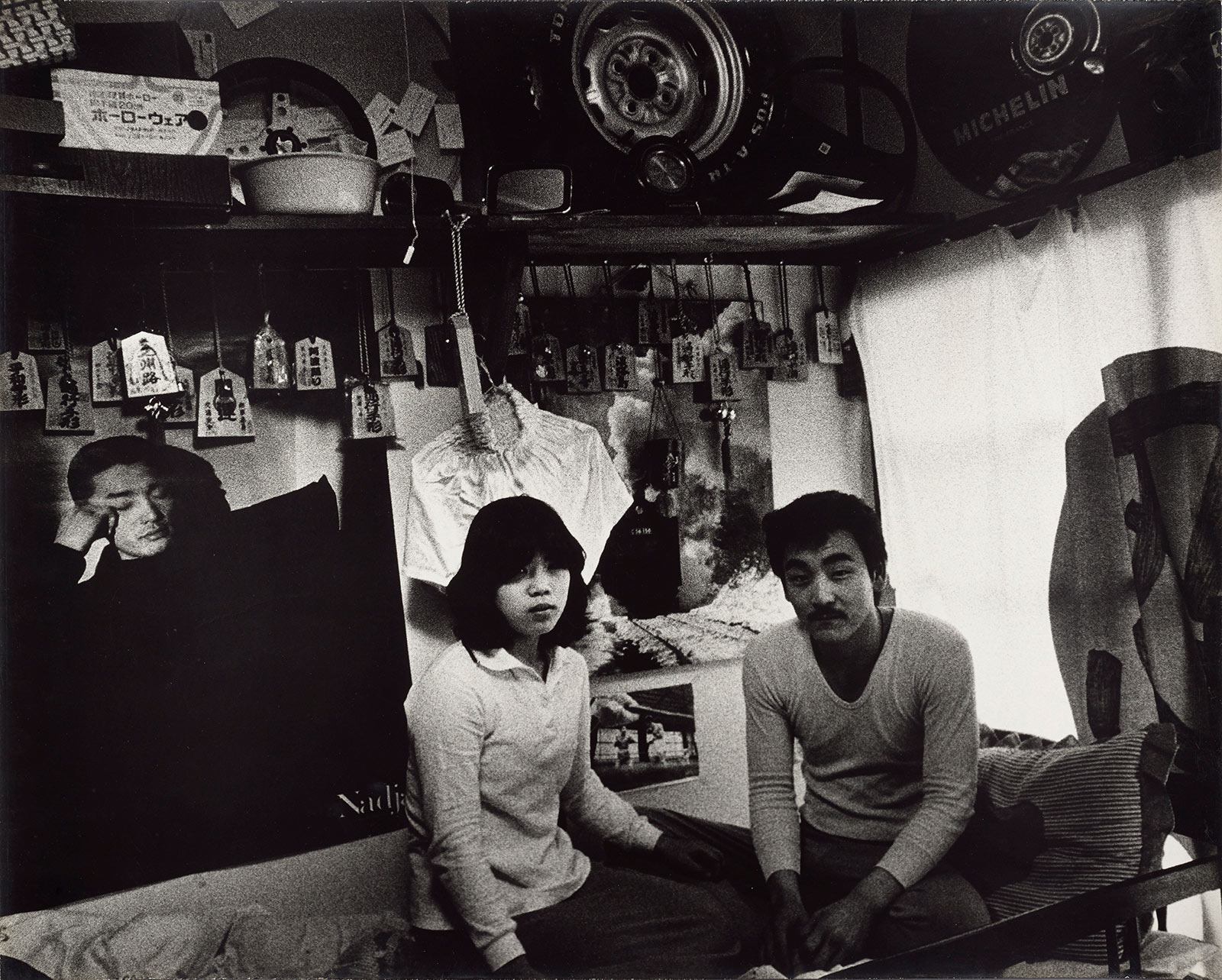 A young Japanese woman and man sit together, facing the camera, in a small apartment.