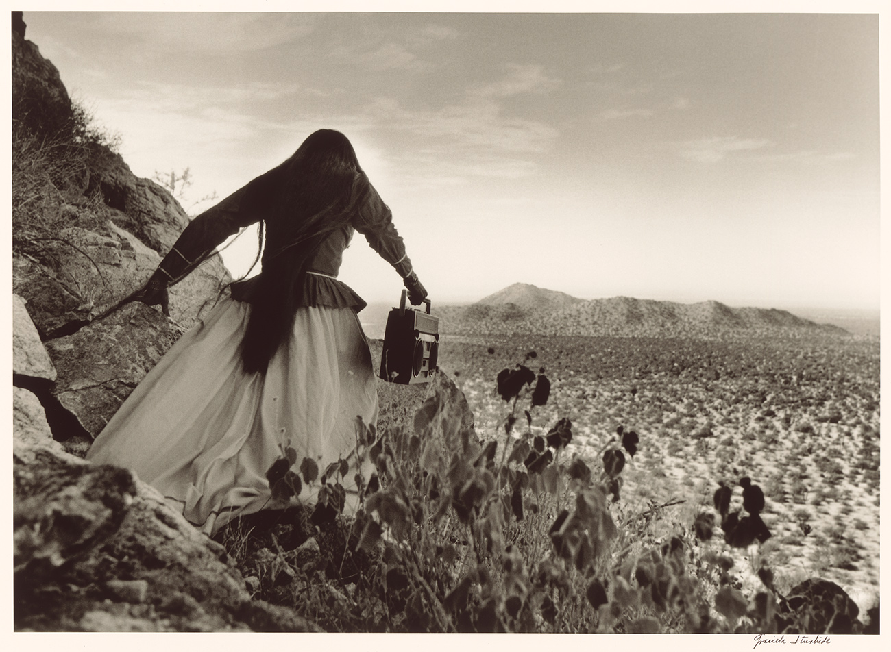 A woman stands looking out on a desert landscape. Her dark hair is long and she wears a big white skirt and dark jacket on top.