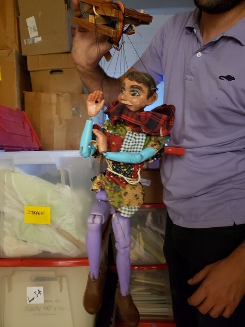 Man holding a marionette puppet, with stacks of boxes and bins in the background