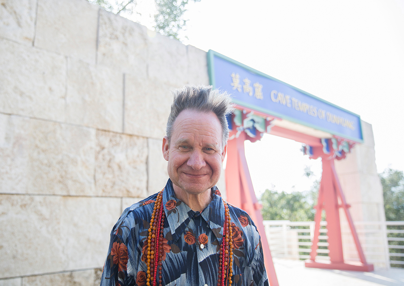 Sellars wears a patterned blue shirt and is standing outside a stone archway featuring a blue sign and red decorative arch at the Getty Center.