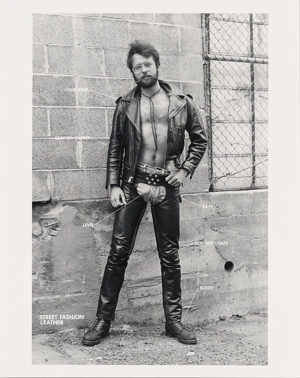 Man with glasses and beard poses in front of a concrete building. He wears a leather jacket and chaps.