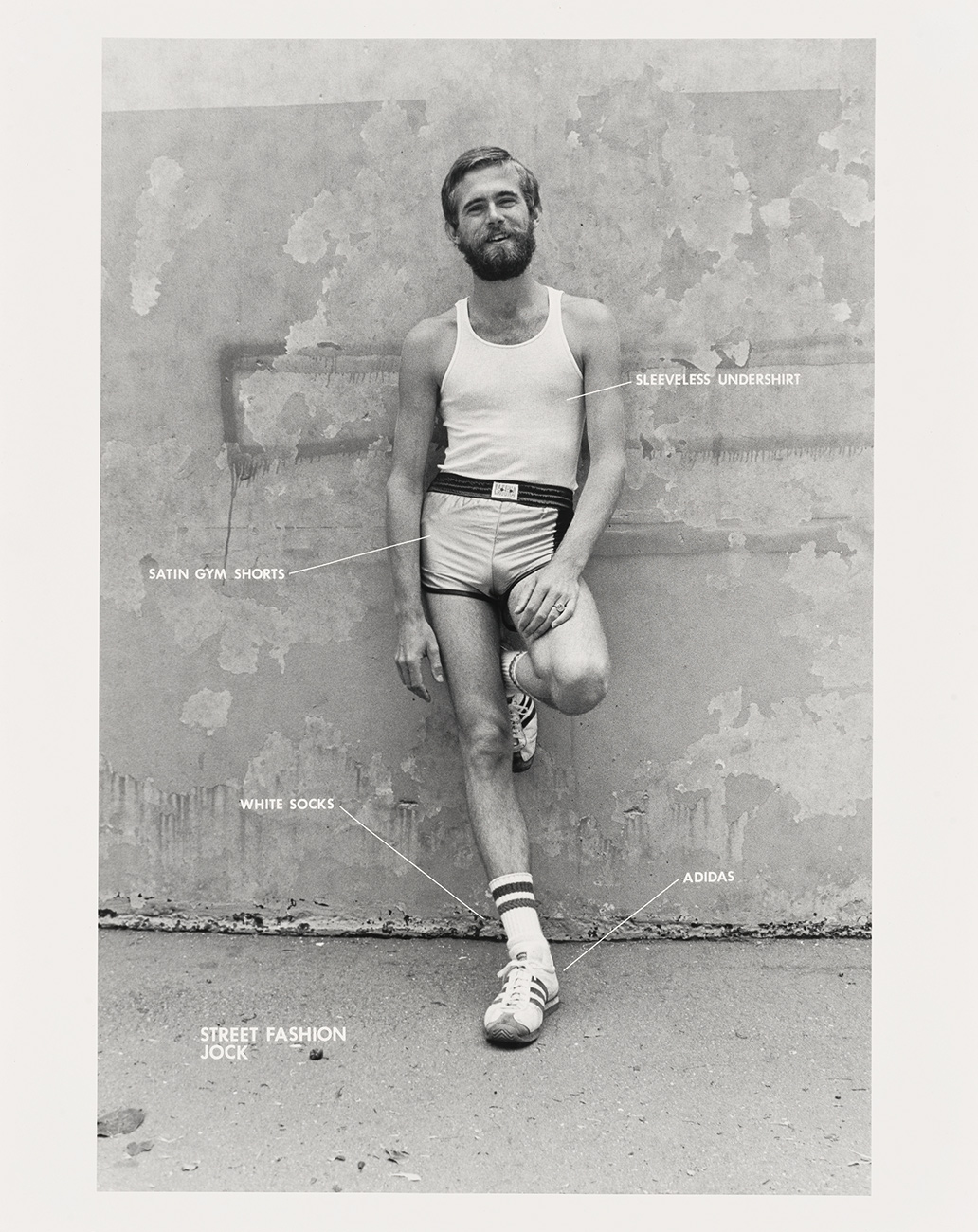 Man poses, leaning against a wall. He wears satin shorts, white socks and a tank top.