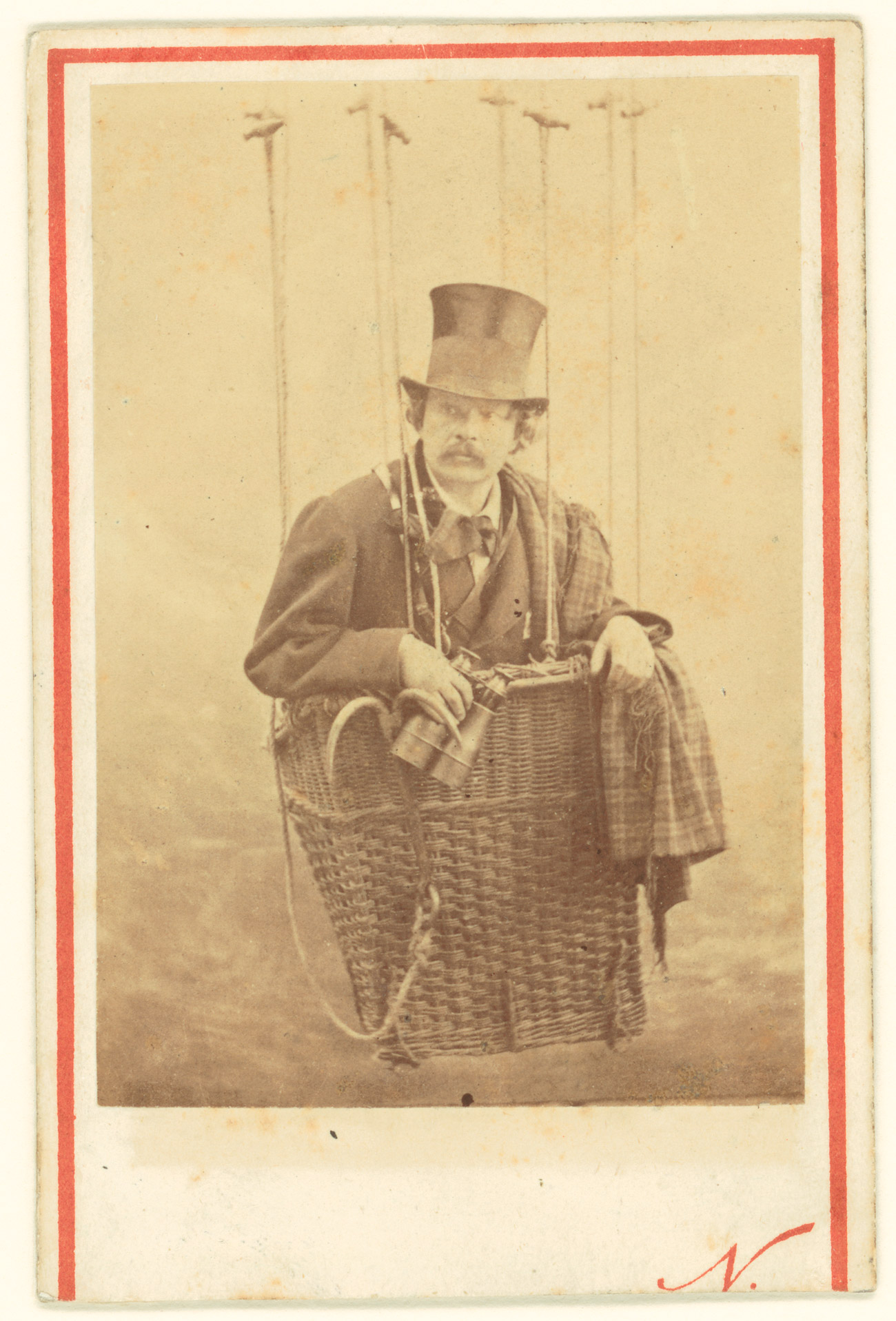 Photograph of a man wearing a top hat and holding binoculars sitting in a small hot air balloon basket
