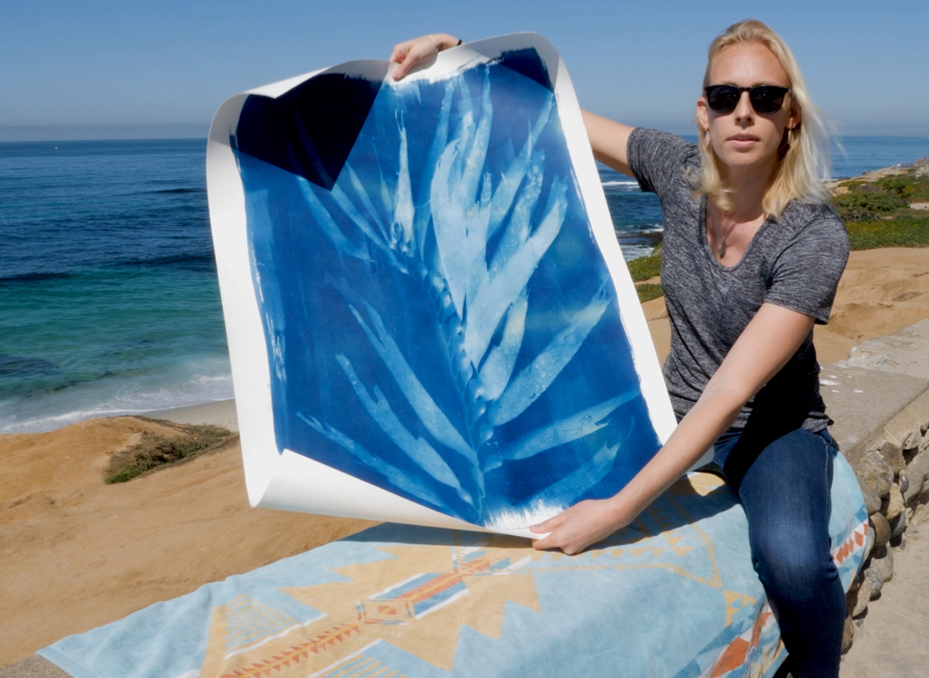 Oriana Poindexter sits on a blanket by the ocean and holds a big print of kelp leaves.