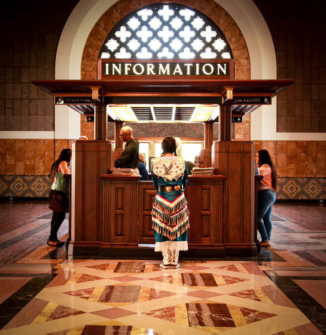Indigenous person wearing blue, red, and white dress with fringe standing at an information desk, in a large hall with tiled floors and grand window