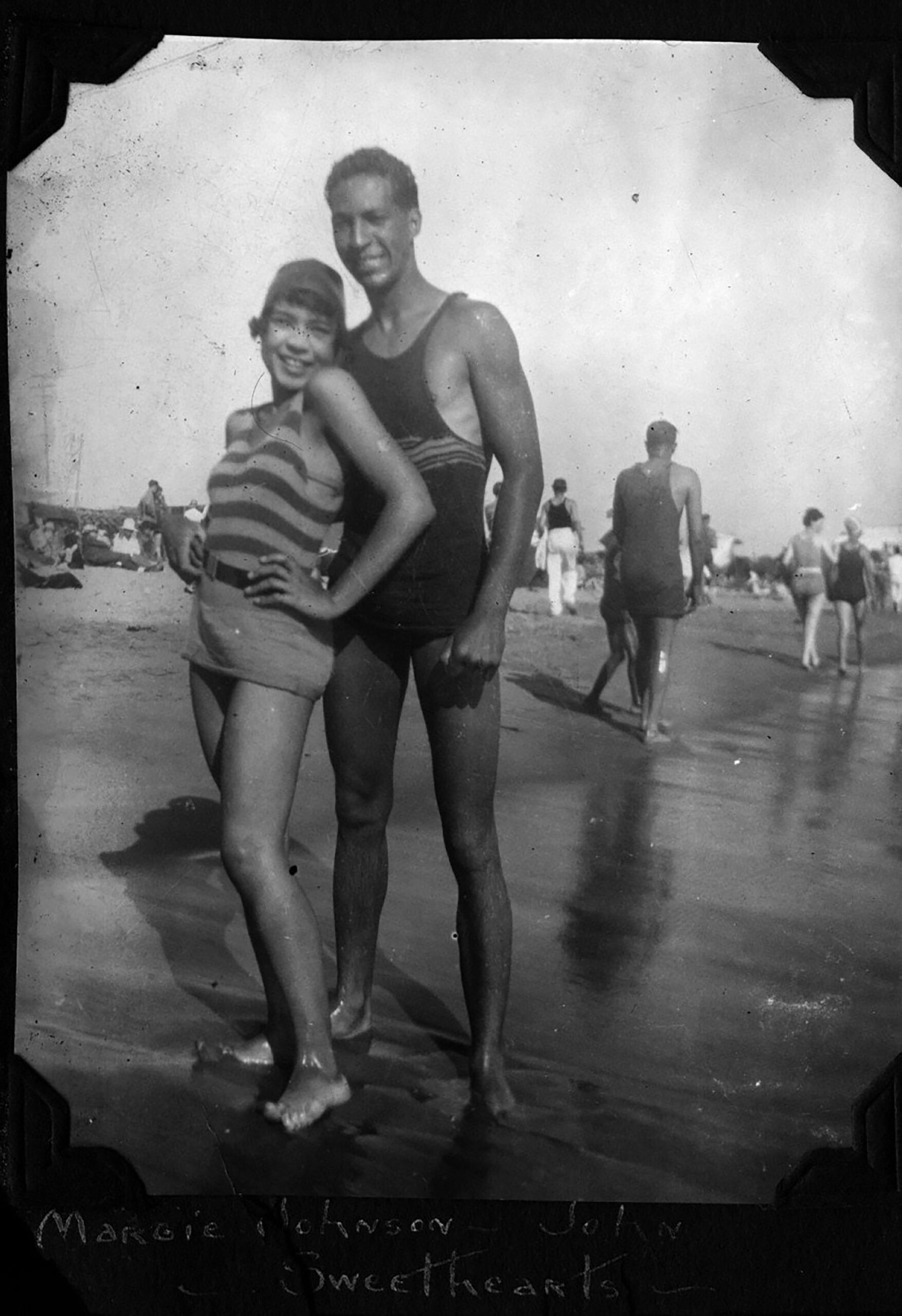 A Day at Bruce’s Beach, Manhattan Beach, California, July 10, 1927. Sweethearts Margie Johnson and John Pettigrew strike a pose at the crowded Pacific Ocean shoreline. Photo: From the private LaVera White Collection of Arthur and Elizabeth Lewis Photograph of African American couple, Margie Johnson and John Pettigrew standing together on Bruce's Beach in Manhattan Beach, California, July 10, 1927